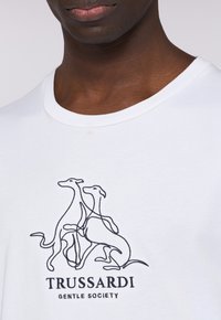 Camisa de algodón blanca con un dibujo a línea negro de dos perros y el texto "TRUSSARDI GENTLE SOCIETY" debajo en letras en negrita.