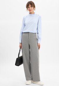 Vrouw in een lichtblauwe gestreepte blouse met een ruches-kraag, grijze wijde broek, witte sneakers, met een zwarte handtas op een witte achtergrond.