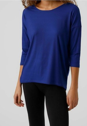 Vrouw draagt een loszittend, geribbeld, blauw shirt met lange mouwen en zwarte broek, staand tegen een effen lichtgrijze achtergrond.