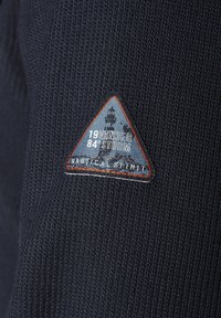 Marineblauer Strickpullover mit einem dreieckigen Stoffpatch, der ein Leuchtturm-Design, den Schriftzug "VANDERSTORM 1984" und den Text "NAUTICAL SPIRIT" in Weiß zeigt.