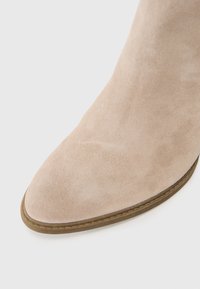 Beige semsket ankelstøvel med spiss tå, lysebrune sømmer og flat såle. Overflaten er glatt og myk å ta på.