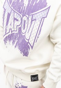 Hvid sweatshirt med et stort lilla "TAPOUT" grafik med et tekstureret design. Inkluderer et sort logomærke nederst til venstre.