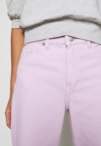 Jeans in denim lilla con taglio dritto, dotati di chiusura con zip e bottone, tasche frontali e leggera texture. Abbinati a una felpa grigia.