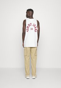 Camiseta sin mangas blanca con el texto rojo "TOMMY JEANS" en la parte de atrás, combinada con pantalones cargo color beige, que presentan bolsillos superpuestos y un ajuste relajado.