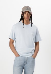 Light blue short-sleeve t-shirt with a round neckline and small embroidered text on the left chest, paired with light blue jeans and a grey cap.
