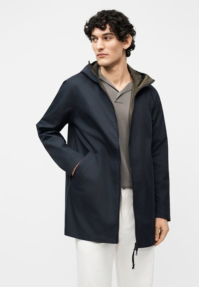 Jeune homme aux cheveux bouclés portant une veste à capuche bleu marine sur un t-shirt gris et un pantalon blanc, les mains dans les poches, regardant sur le côté.