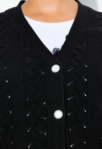 Černý pletený kardigan s výstřihem do V, složitým krajkovým vzorem a stříbrným knoflíkem se středem připomínajícím bílou perlu.