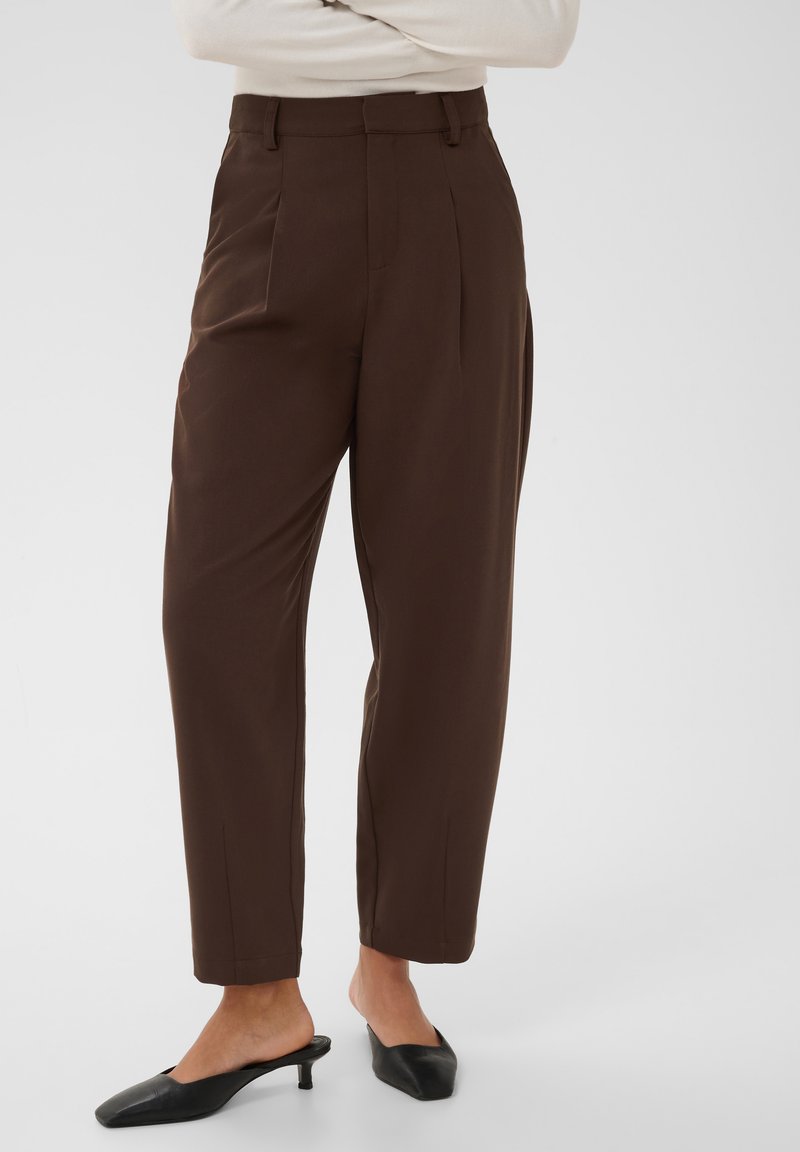 Femme portant un pantalon marron taille haute et fuselé, et des mules noires à talons bas, debout les bras croisés.