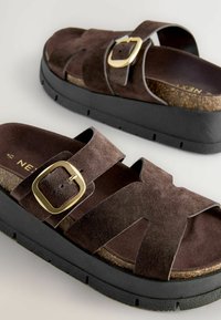 Sandale din suede maro cu o talpă din plută, dotate cu bretele intersectate și un detaliu cu cataramă aurie, așezate pe o talpă groasă neagră.