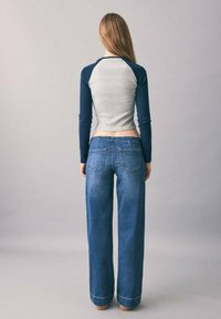 Una giovane donna sta in piedi girata di spalle, indossando una maglietta a maniche lunghe grigia e blu navy e un paio di jeans blu larghi, su uno sfondo semplice.