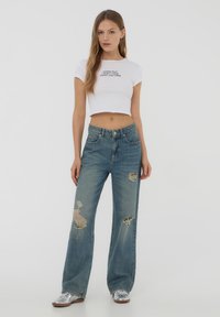 T-shirt bianca a maniche corte con testo nero, abbinata a jeans a vita alta blu chiaro con dettagli strappati e bordi sfilacciati. Sneakers argento.