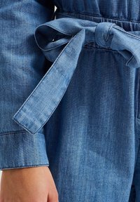 Tuta in denim con cintura, di colore blu medio, texture morbida e silhouette aderente con maniche a sbuffo.