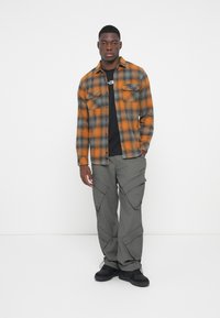 Camisa de franela en cuadros naranjas y negros, combinada con pantalones cargo gris oscuro con múltiples bolsillos y deportivas negras.