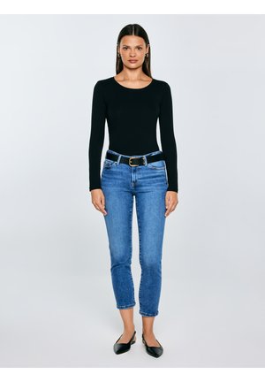 Vrouw staat recht vooruit kijkend, gekleed in een zwart top met lange mouwen, blauwe cropped spijkerbroek met zwarte riem, en zwarte puntige slingback platte schoenen.