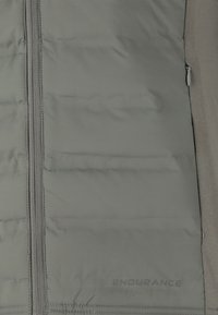 Chaleco acolchado en gris claro con textura de acolchado, cremallera frontal y un sutil logotipo "ENDURANCE" en la esquina inferior derecha.