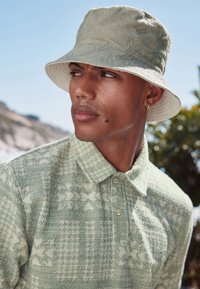 Giovane uomo che indossa un cappello a secchiello beige a motivi e un maglione verde lavorato a maglia con disegni geometrici, che guarda di lato all'aperto in una giornata di sole.