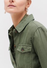 Veste olive en tissu durable, dotée d'un col pointu, d'une fermeture à boutons sur le devant et de deux poches poitrine à boutons.