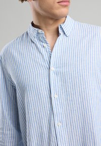 Young man wearing a blue and white vertically striped button-up shirt with an open collar, facing slightly to the side.