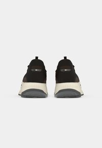 Sapatos atléticos pretos e castanhos com uma sola almofadada bege, apresentando um cabedal em tecido de malha, alça de puxar e detalhes discretos do logótipo.