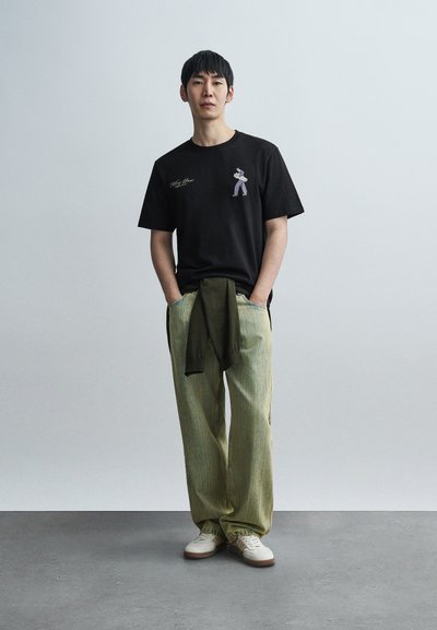 Μαύρο βαμβακερό μπλουζάκι με μικρό μωβ γραφικό, σε συνδυασμό με ανοιχτό πράσινο, φαρδιά ριγέ παντελόνια και λευκά αθλητικά παπούτσια.