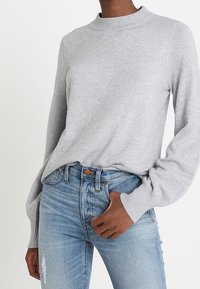 Hellgrauer Strickpullover mit hohem Kragen und langen Ärmeln, kombiniert mit hellblauen, stark verwaschenen High-Waisted-Jeans.