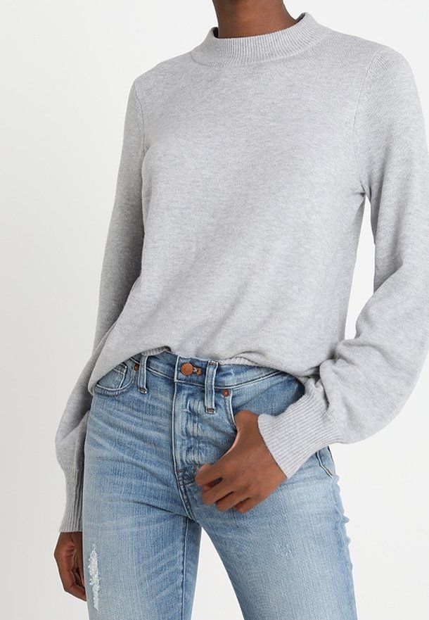 Hellgrauer Strickpullover mit hohem Kragen und langen Ärmeln, kombiniert mit hellblauen, stark verwaschenen High-Waisted-Jeans.
