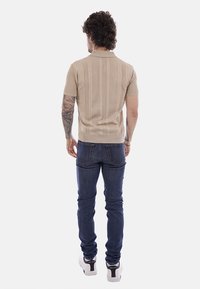 Uomo tatuato con indosso una camicia beige a maniche corte, jeans blu scuro e sneakers bianche, in piedi di spalle contro uno sfondo bianco.
