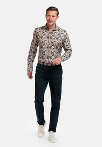 Chemise à manches longues avec un motif floral multicolore, col boutonné, portée avec un pantalon sombre et des baskets beiges. Tissu lisse, design ajusté.