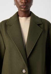 Olive green wool blend coat with a notched collar, single button closure, and soft texture, layered over a beige ribbed sweater.