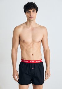 Boxers noirs avec une ceinture élastique rouge arborant le logo "HUGO". Comprend un bouton à l'avant et un détail de couture contrastante.
