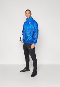 Giacca a vento blu con un design a fantasia, chiusura con zip e orlo elasticizzato, abbinata a pantaloni jogger neri a sigaretta e scarpe nere, mentre tiene un pallone da calcio.