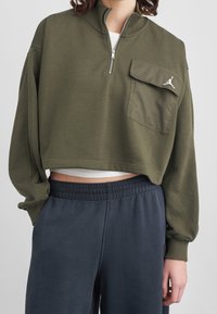 Personne portant un sweat zippé court couleur olive avec une poche poitrine et un logo blanc, un pantalon de survêtement bleu marine et un sous-vêtement blanc, debout.
