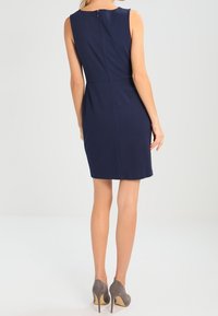 Granatowa sukienka bez rękawów o dopasowanym kroju, z zamkiem z tyłu i długości do kolan, połączona z szarymi szpilkami.
