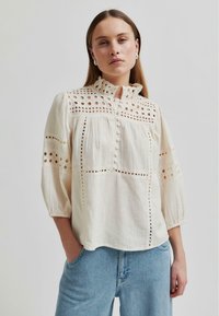 Helle, strukturierte Bluse mit hohem, gerafftem Kragen, Lochstickdetails und Knopfverschluss. Sie hat Puffärmel und eine lockere Passform.