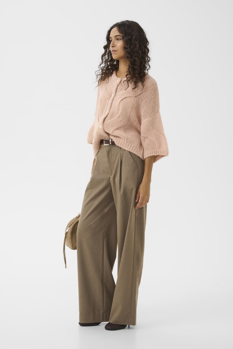 Vrouw met krullend haar, gekleed in een roze chunky brei cardigan en wijde bruine broeken, met een beige tas in haar hand, staand tegen een witte achtergrond.