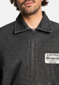 Black denim jacket with a zip front, a pointed collar, and a patch featuring "DC Shoe Company Worldwide Goods Since 1994."