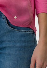 Top rosa a maniche corte realizzato in un tessuto leggero e trasparente, con un bottone decorativo. Abbinato a jeans in denim blu con una texture liscia.