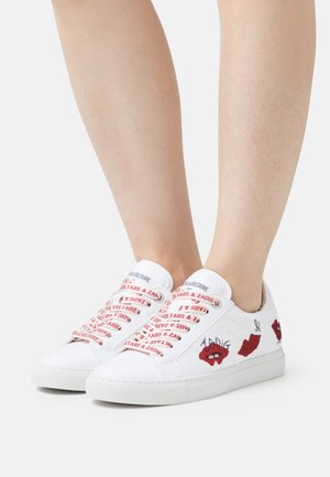 Weiße Low-Top-Sneaker mit roten Lippen und aufgesticktem Text, getragen an nackten Beinen vor einem einfarbigen hellen Hintergrund.