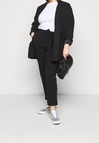 Svart kavaj över en vit topp, svarta byxor med ett bälte, grå sneakers med vita snören, håller en svart texturerad väska.