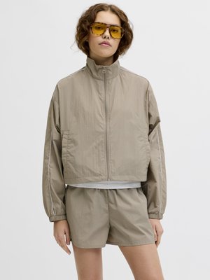 Persona che indossa una giacca beige con cerniera e pantaloncini abbinati, con occhiali da sole a lente gialla, in piedi contro uno sfondo chiaro e uniforme.