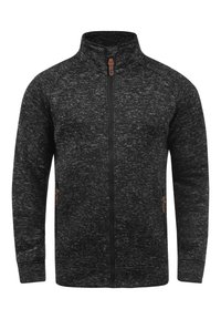 Musta, vetoketjullinen takki, joka on valmistettu teksturoidusta kankaasta, jossa on kirjoluonnoskuvio. Siinä on sivutaskut ja korkea kaulus.