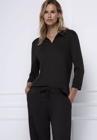 Schwarzes Oberteil mit Reißverschluss, Kragen und dreiviertel Ärmeln, kombiniert mit schwarzen Hos̈n mit Kordelzug, beide aus glattem Material und lockerem Schnitt.