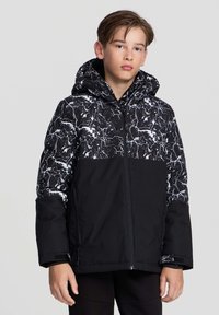 Nastoletni chłopak w czarnej i białej, marmurkowej kurtce z kapturem, stoi z neutralnym wyrazem twarzy na jednolitym szarym tle.