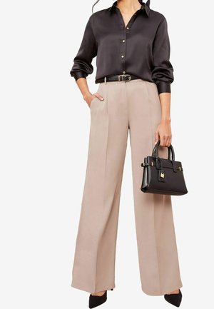 Vrouw draagt een zwarte zijden blouse met knopen, gestoken in beige broek met wijde pijpen, zwarte riem, zwarte schoenen met een puntige neus, en draagt een zwarte handtas.