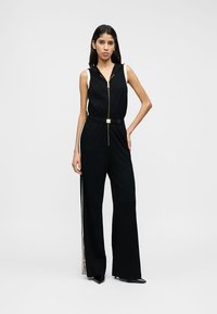 Black jumpsuit with sleeveless design, gold zipper, and belted waist. Features side stripe detail and wide-leg cut. Smooth texture.
