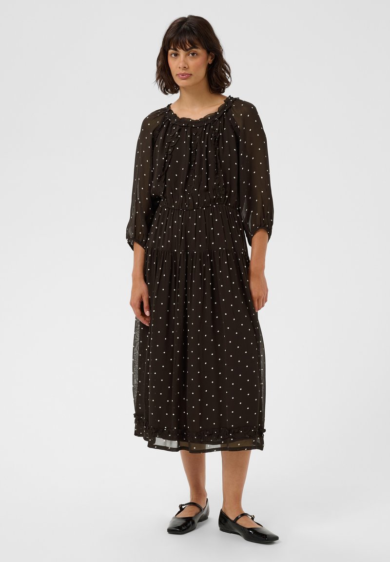 Femme debout portant une robe noire mi-longue à pois blancs, un col froncé et des chaussures plates noires avec des brides à la cheville.