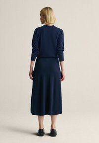 Laivastonsininen neulemekko, jossa on pitkät hihat, laskeutuva hame ja pyöreä pääntie, valmistettu pehmeästä kankaasta. Mustat kengät täydentävät asun.