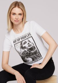 Biała koszulka z czarno-białym nadrukiem graficznym oraz napisem. Krótkie rękawy, okrągły dekolt, miękki bawełniany materiał. Czarne spodnie.