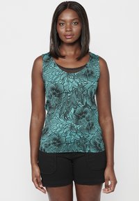 Débardeur sans manches en teal avec motif floral noir ; il présente un col rond et un bord noir texturé au niveau du col. Associé avec un short noir.