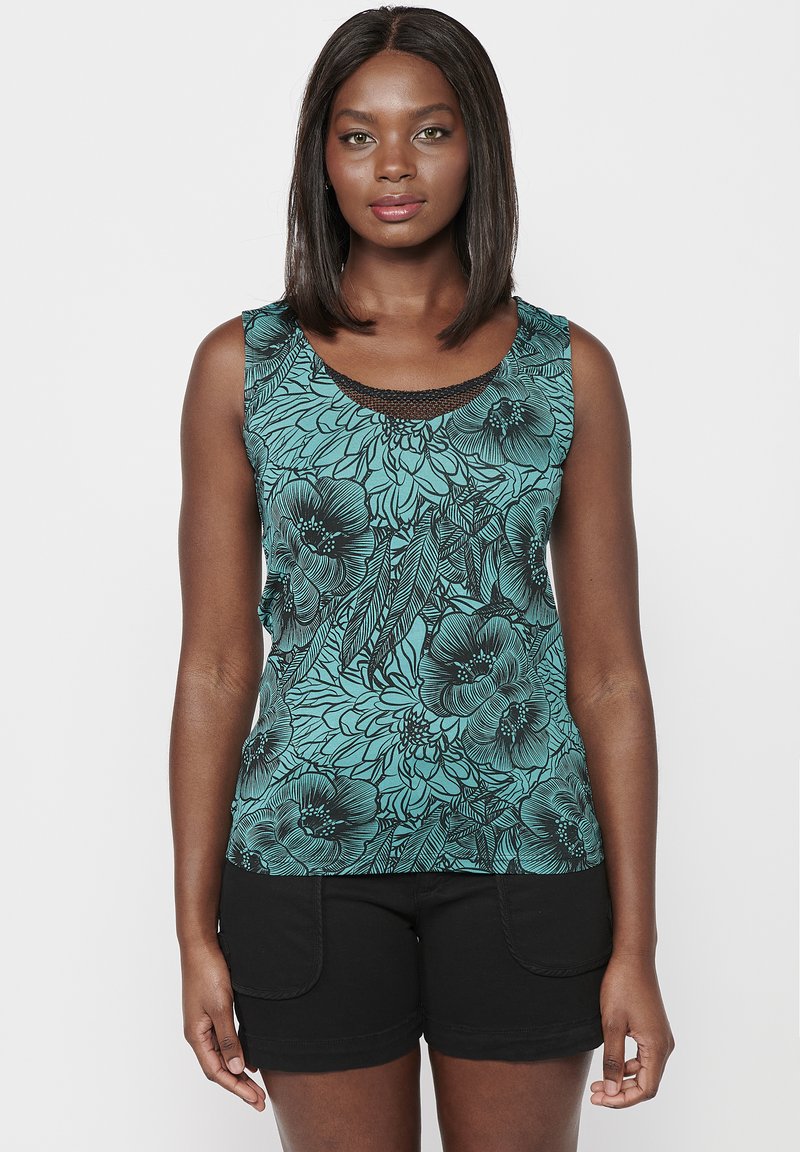 Débardeur sans manches en teal avec motif floral noir ; il présente un col rond et un bord noir texturé au niveau du col. Associé avec un short noir.